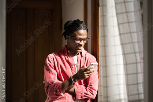 Photography Smiling young black man holding cellphone, standing near window