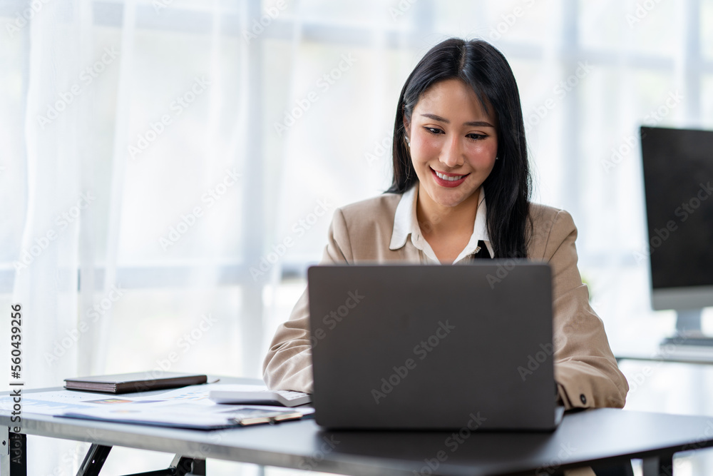 Asian young woman businesswoman using a laptop sitting smiling using modern analytical concepts While happily working at the desk in the office. Asian young woman businesswoman using a laptop sitting smiling using modern analytical concepts While happily working at the desk in the office.