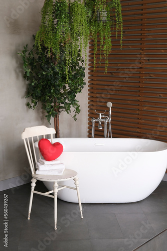 Modern bathroom interior design with concrete walls, wooden slats and white bathtub. vintage chair with towels and heart. Valentine's day concept, relaxation, massage, gifts, care, rest.