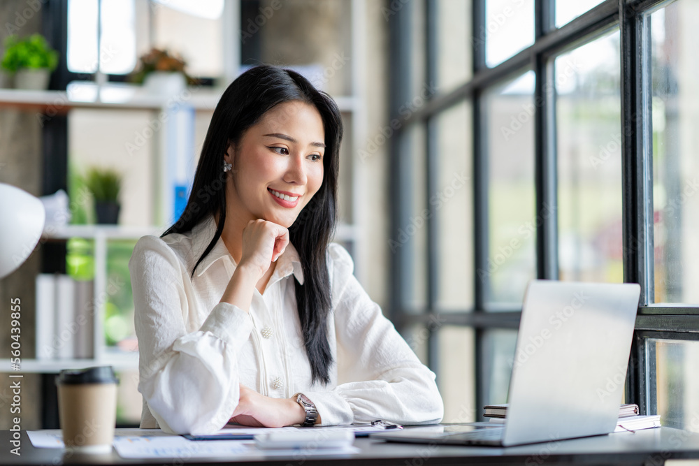Fototapeta premium Portrait of a beautiful cheerful Asian businesswoman sitting at her desk with a cup of coffee while taking a break with a happy smile in her upcoming successful business.