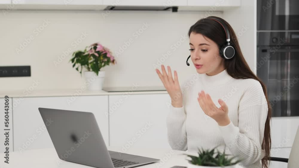 Female employee in headset talks to colleagues at online meeting via Notebook. Brunette woman explains project details at kitchen table slow motion Female employee in headset talks to colleagues at online meeting via Notebook. Brunette woman explains project details at kitchen table slow motion