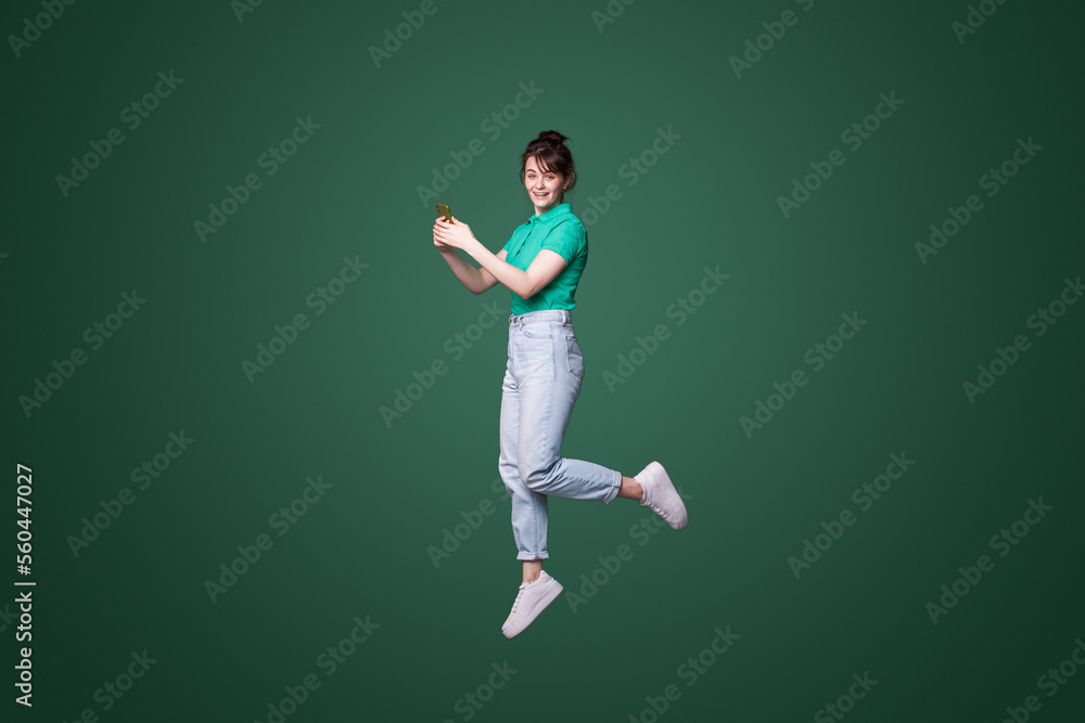 Full body portrait of young smiling happy woman jump high hold in hand mobile cell phone isolated on green color background. People lifestyle concept Full body portrait of young smiling happy woman jump high hold in hand mobile cell phone isolated on green color background. People lifestyle concept