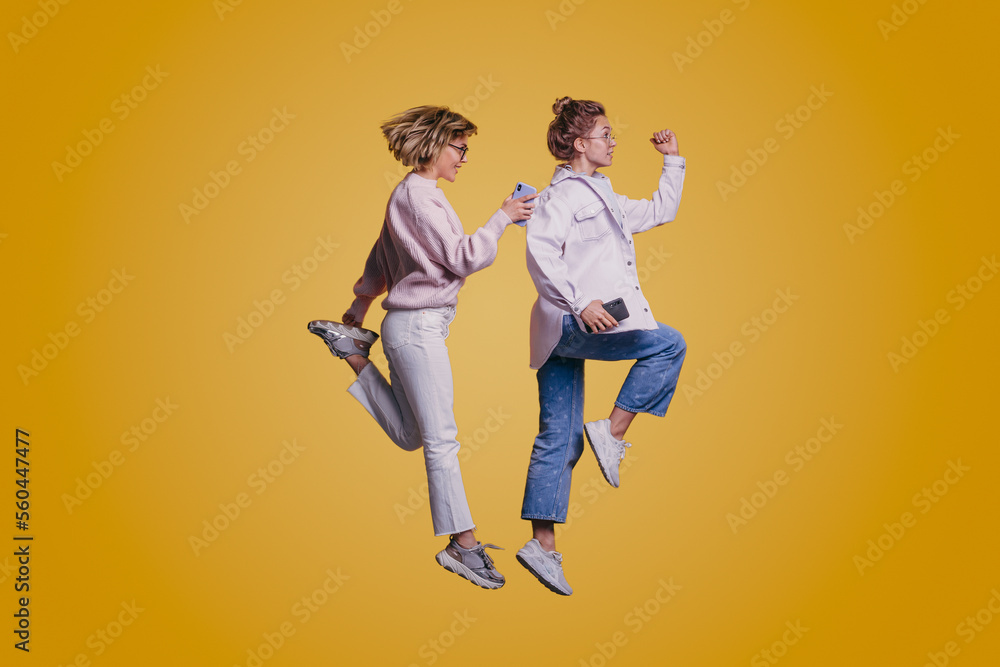 Full length body size view of two cheerful women jumping holding their phones isolated on yellow color background Full length body size view of two cheerful women jumping holding their phones isolated on yellow color background