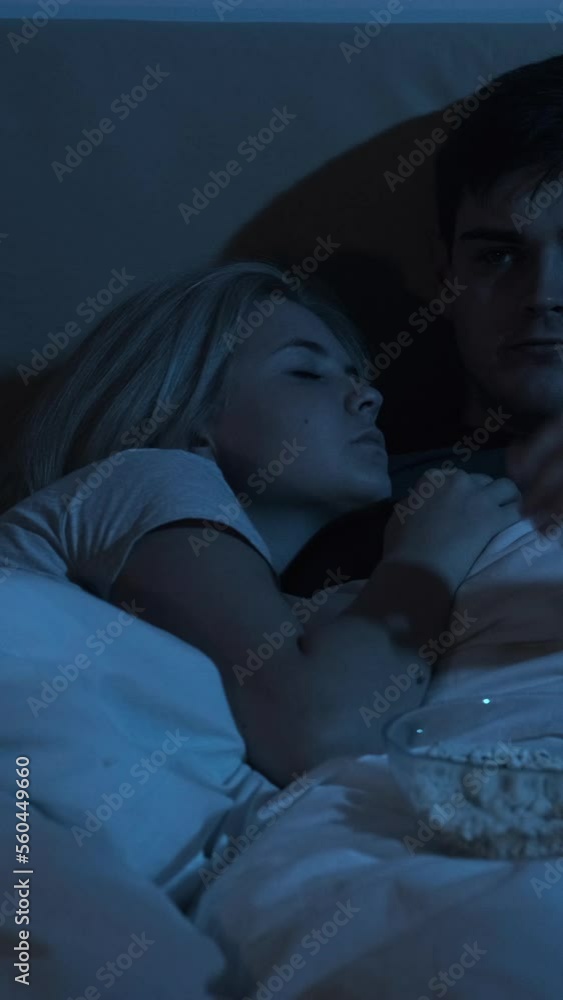 Night movie. Insomnia problem. Late home leisure. Bored restless husband watching TV eating popcorn in bed with tired wife sleeping in dark bedroom. Vertical video. Night movie. Insomnia problem. Late home leisure. Bored restless husband watching TV eating popcorn in bed with tired wife sleeping in dark bedroom. Vertical video.