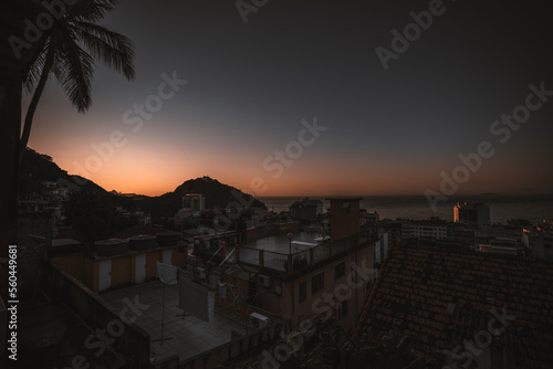 A wide-angle sunset shot from a rooftop in a house placed in a Brazilian favela in Rio, overlooking the calming sea and the rooftops of the surrounding houses with visible wear and tear of poverty