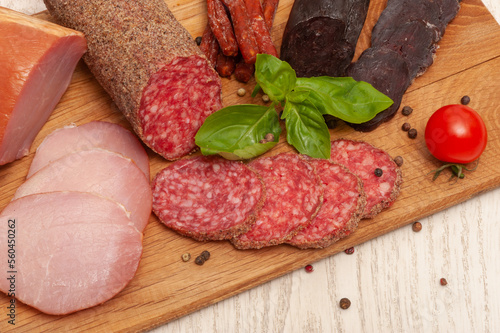 Different types of sausages ham, mahan, salami on a wooden board decorated with tomatoes and basil