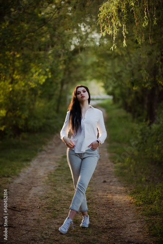 Fototapeta Naklejka Na Ścianę i Meble -  Sexy sensual brunette walks in a summer forest at sunset in a white blouse and jeans