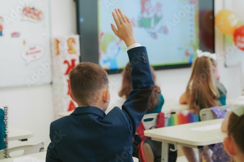 children in school uniforms sitting by desks. Boy raising hand to answer