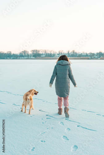 Winter walk of young beautiful brunette woman wearing warm clothes in snow with dog golden. Snowing winter concept.