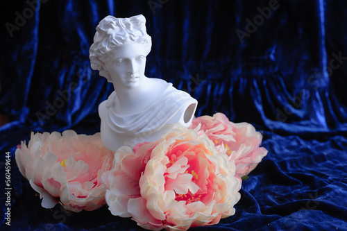 Greek sculpture among pink peonies on a blue velvet background. Copy space