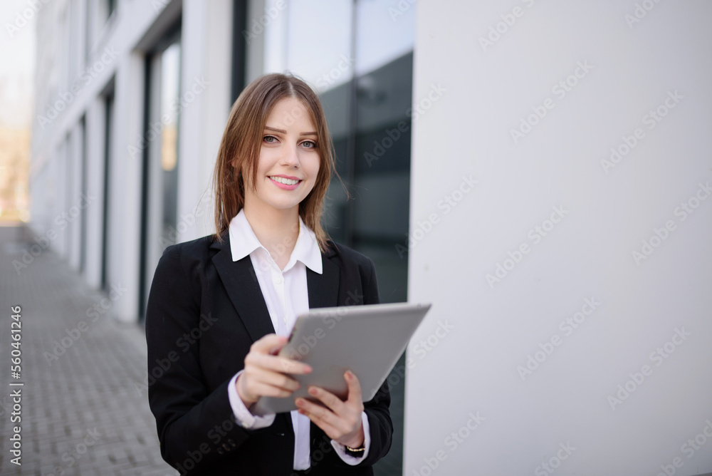 A woman manager in a white shirt stands in front of a glass modern corporation business building, holding a tablet in her hands, browsing the web, answering emails. Copy space. A woman manager in a white shirt stands in front of a glass modern corporation business building, holding a tablet in her hands, browsing the web, answering emails. Copy space.