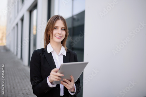  A woman manager in a white shirt stands in front of a glass modern corporation business building, holding a tablet in her hands, browsing the web, answering emails. Copy space.
