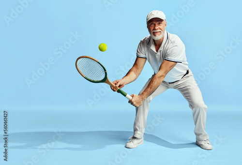 Active retirement life. Portrait of handsome senior man in stylish white outfit playing tennis over blue background. Concept of leisure activity, hobby, lifestyle © master1305
