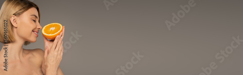 side view of cheerful woman with closed eyes smelling orange isolated on grey, banner.