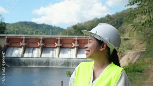 Smiling female engineer in green vest and helmet standing outside against background of hydroelectric dam, floodgate with flowing water through gate.