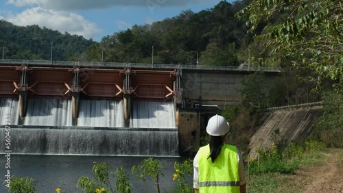 Female engineer wearing green vest and white helmet working outside dam with hydroelectric power plant and irrigation. Renewable energy system. Sustainable energy concept.