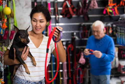 Asian woman holding her little dog in hands while choosing new accessories in salesroom of pet shop. Elderly man making purchases in background.