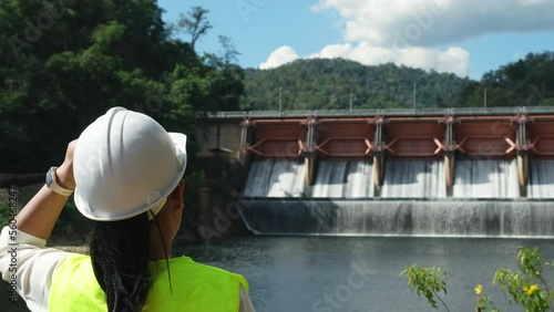 Female engineer wearing green vest and white helmet working outside dam with hydroelectric power plant and irrigation. Renewable energy system. Sustainable energy concept.