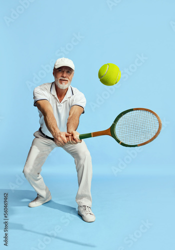 Serving ball. Portrait of handsome senior man in stylish white outfit playing tennis over blue background. Concept of leisure activity, hobby, lifestyle © master1305