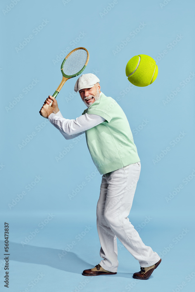 Happy. Portrait of handsome senior man in stylish outfit playing tennis, serving ball over blue background. Concept of leisure activity, hobby, lifestyle