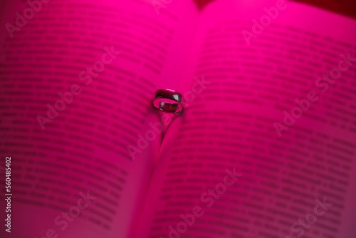 Wedding ring in open book and heart-shaped shadow. Pink neon light illuminates pages. Concept of Valentine's Day. Top view and text not readable due to blurring. Eengagement proposal. photo