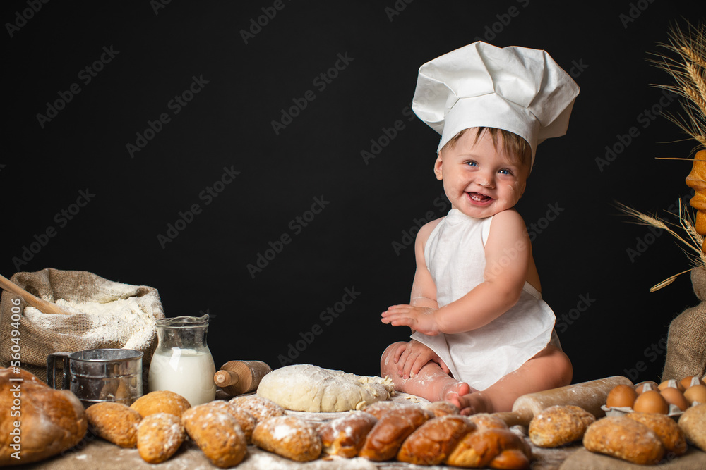 Funny Little boy sits in cook costume among baking bread rolls on the table and kneading the dough Funny Little boy sits in cook costume among baking bread rolls on the table and kneading the dough