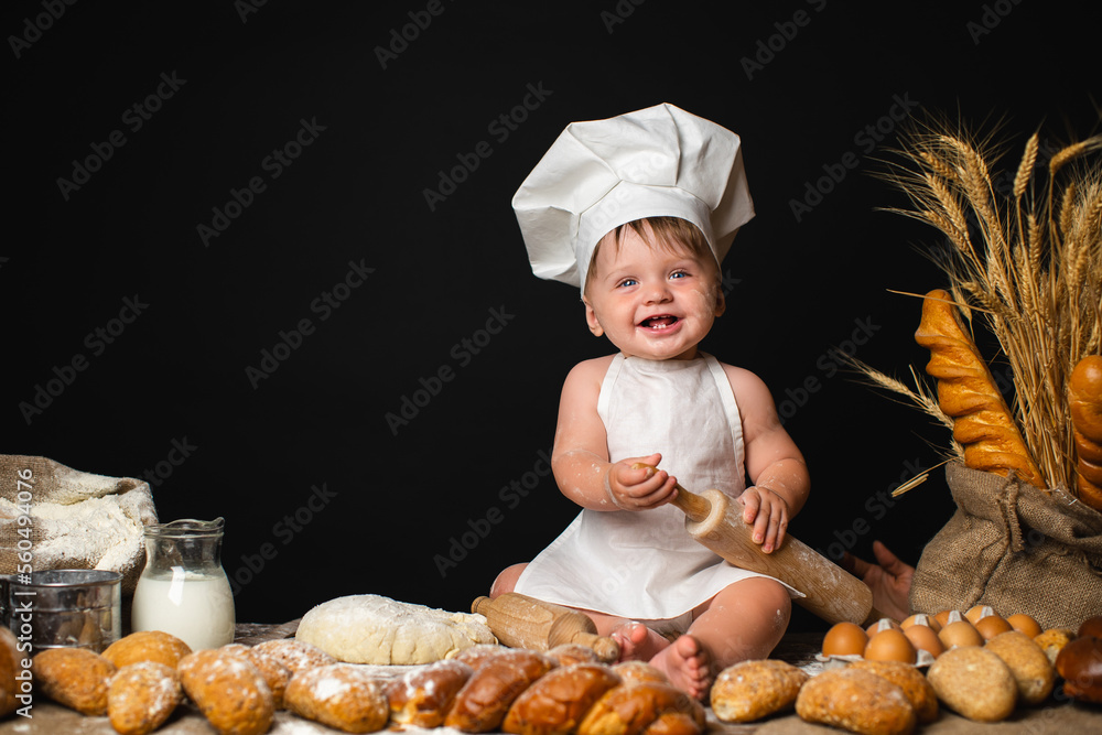 Laughing little boy sits in cook costume among baking bread rolls on the table on black background Laughing little boy sits in cook costume among baking bread rolls on the table on black background