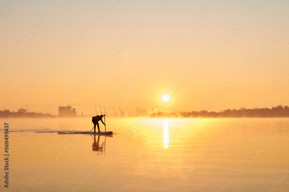 Silhouette of man rowing on SUP (stand up paddle board) at sunrise in a foggy haze in the Danube river at cold season