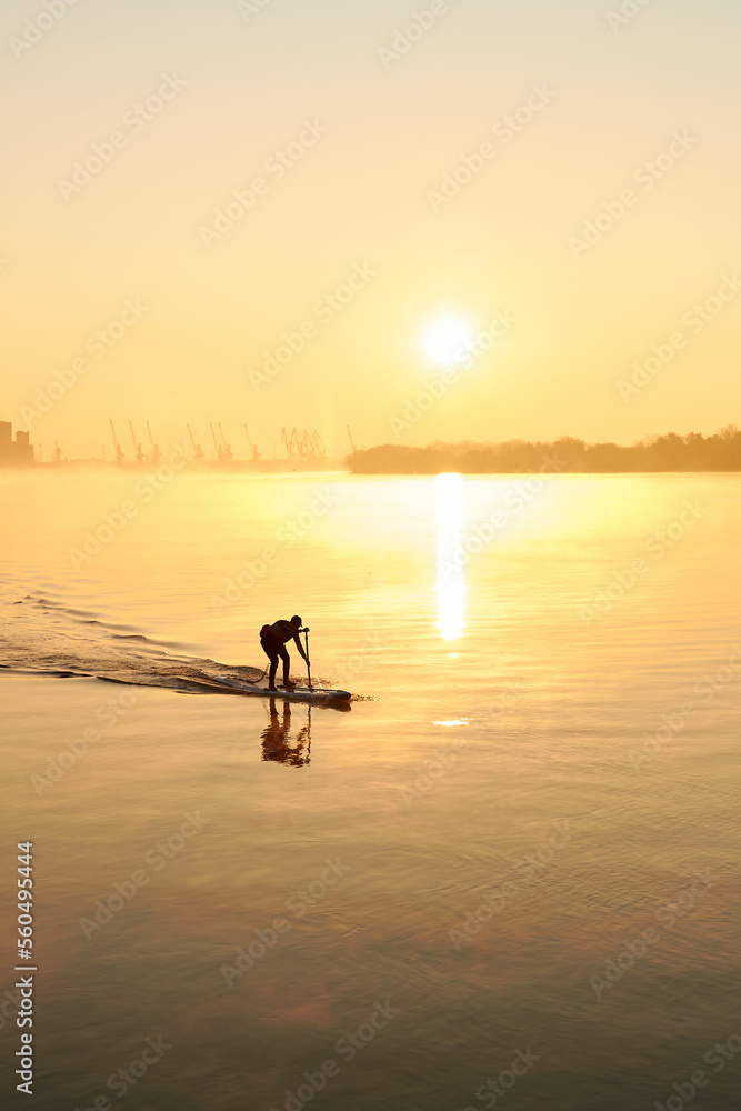Silhouette of man rowing on SUP (stand up paddle board) at sunrise in a foggy haze in the Danube river at cold season