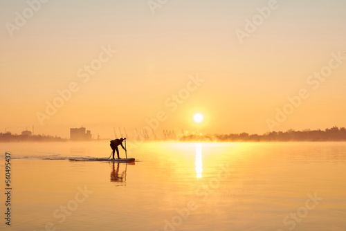 Silhouette of man rowing on SUP (stand up paddle board) at sunrise in a foggy haze in the Danube river at cold season © watcherfox