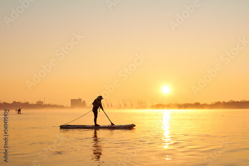 Silhouette of woman standing on SUP (stand up paddle board) at sunrise in a foggy haze in the Danube river at cold season