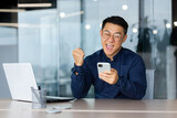 A happy young man, an Asian businessman, a student sits in the office at a table with a laptop, uses a mobile phone. Rejoices, celebrates, received good news, win, shows with hand yes