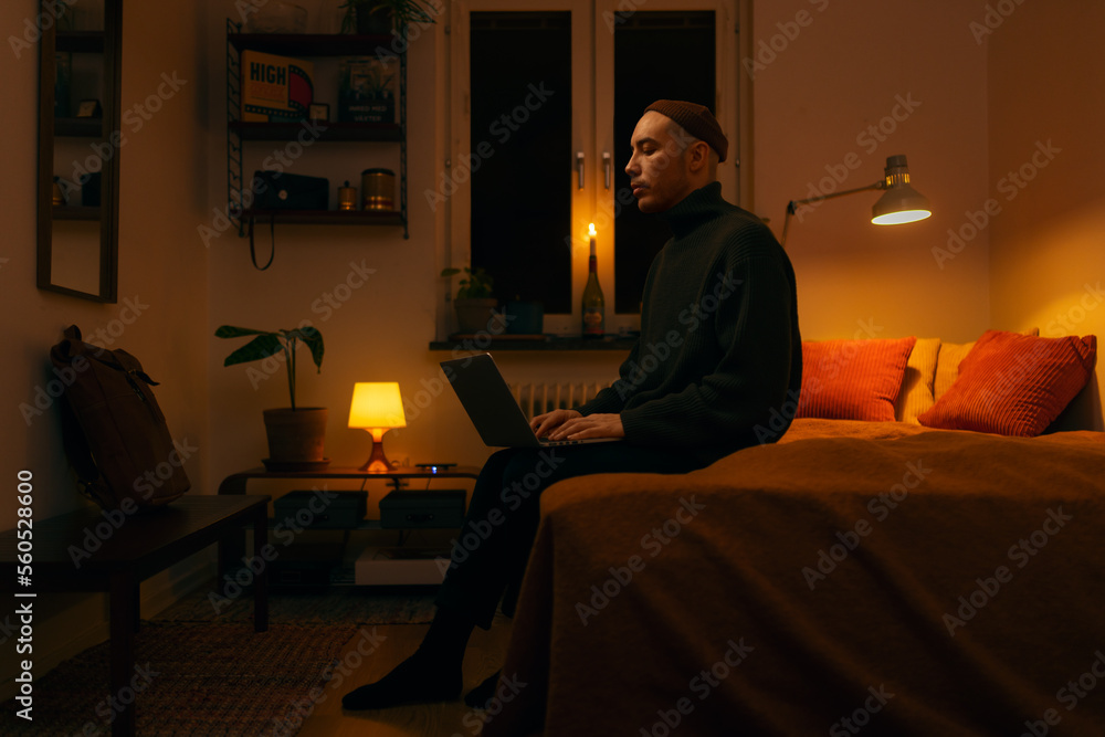 A caucasian man sitting on a bed in a bedroom working on a laptop in the evening lit by a warm light.