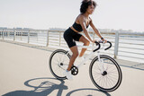 A curly-haired sports workout woman dressed in a black tracksuit poses with a white bicycle on the street