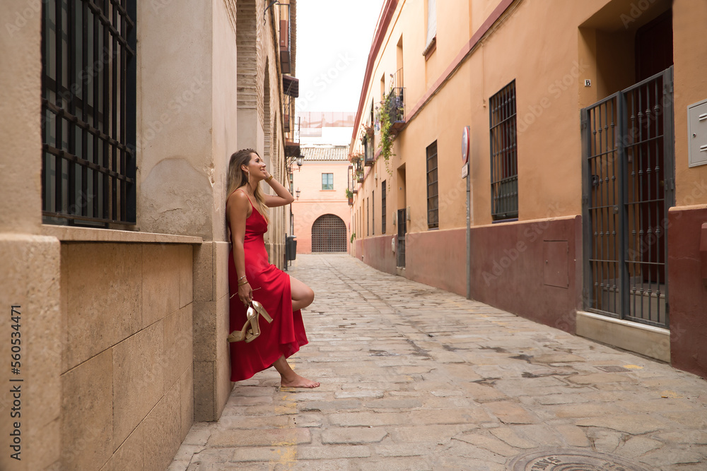 Young, attractive, blonde woman, wearing an elegant red party dress and holding golden high heels in her hand, barefoot, leaning against the wall of a city alley. Concept beauty, fashion, elegance.