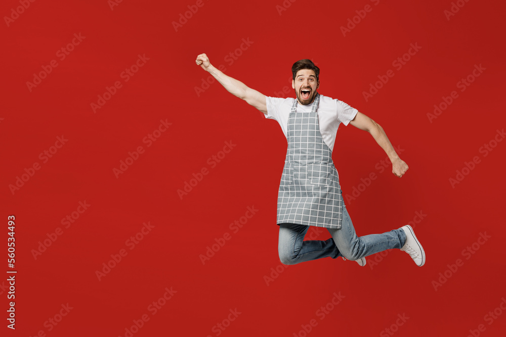 Full body young happy male housewife housekeeper chef cook baker man wear grey apron jump high clench fist pov super hero flying gesture isolated on plain red background studio. Cooking food concept.