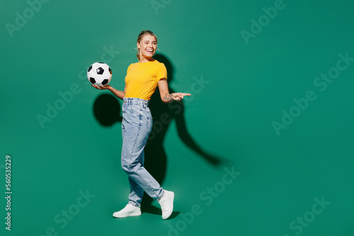 Fototapeta Naklejka Na Ścianę i Meble -  Full body fun young woman fan wear yellow t-shirt cheer up support football sport team hold in hand soccer ball watch tv live stream point index finger aside on area isolated on dark green background.