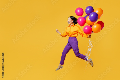 Full body side profile view happy fun young woman wear casual clothes celebrating hold bunch of balloons jumping high run fast isolated on plain yellow background. Birthday 8 14 holiday party concept.
