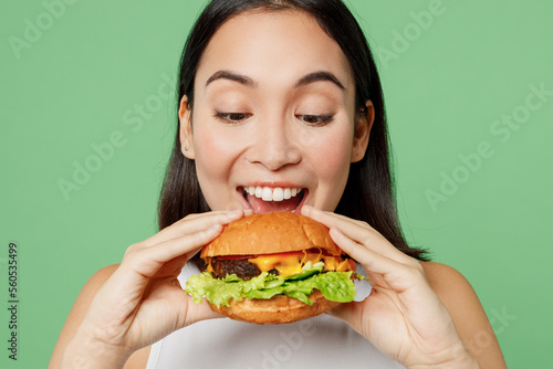 Close up young happy cheerful woman wear white clothes holding eating biting tasty burger isolated on plain pastel light green background. Proper nutrition healthy fast food unhealthy choice concept.