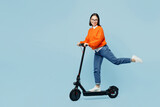 Full body side view smiling happy young woman of Asian ethnicity in orange sweater glasses riding electric scooter isolated on plain pastel light blue cyan background studio People lifestyle concept
