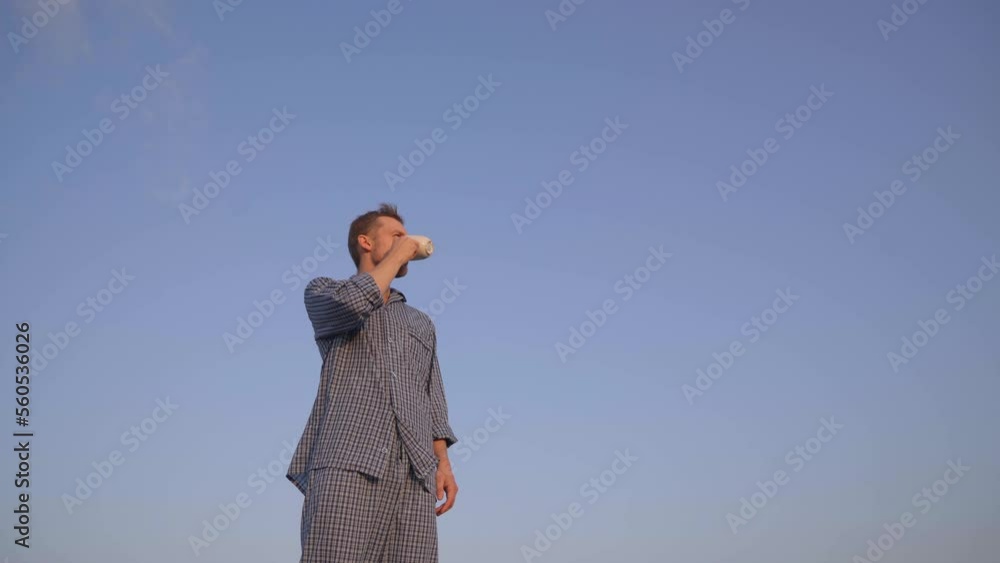 Adorable caucasian male standing outdoor in nightwear looking forward, drinking coffee or tea. Clear blue sky at background. High quality 4k video footage Adorable caucasian male standing outdoor in nightwear looking forward, drinking coffee or tea. Clear blue sky at background. High quality 4k video footage
