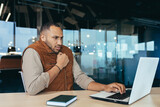 African american man freezing in office, no heating in workplace, man in outerwear sitting at desk using laptop at work.