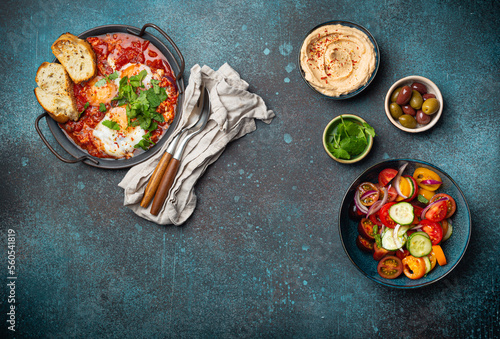 Middle Eastern traditional breakfast or brunch with eggs Shakshouka in pan with toasts, fresh vegetables salad, hummus and olives on rustic concrete background table from above, space for text