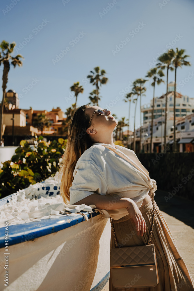 A girl in sunglasses and a long skirt on a summer vacation. A woman against the background of palm trees and a warm city, a tourist rests near a boat. Summer photos.