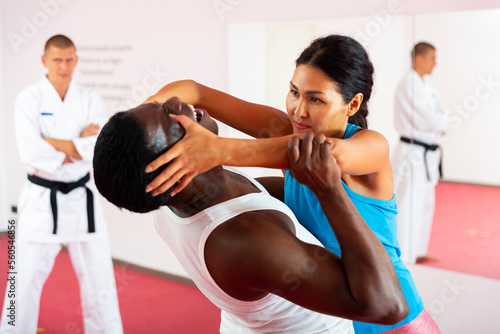 Asian woman exercising eye-gouging move with African-american man during self-protection training, Caucasian man in kimono standing and watching.