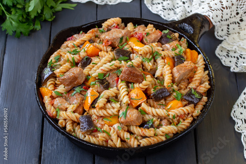 One skillet pork and sauteed vegetables. One Pot pasta dish recipe loaded with fried pork meat, tomatoes, yellow bell pepper, aubergine and onion. Dark wooden table, selective focus, horizontal.