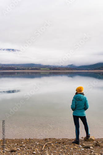 Woman wearing a jacket with her hands in her pockets looking the fog on the mountain in the other side of the lake