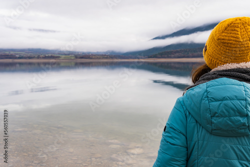 Rear view of woman wearing warm clothes enjoying amazing view of the lake and the mountains with fog the other side
