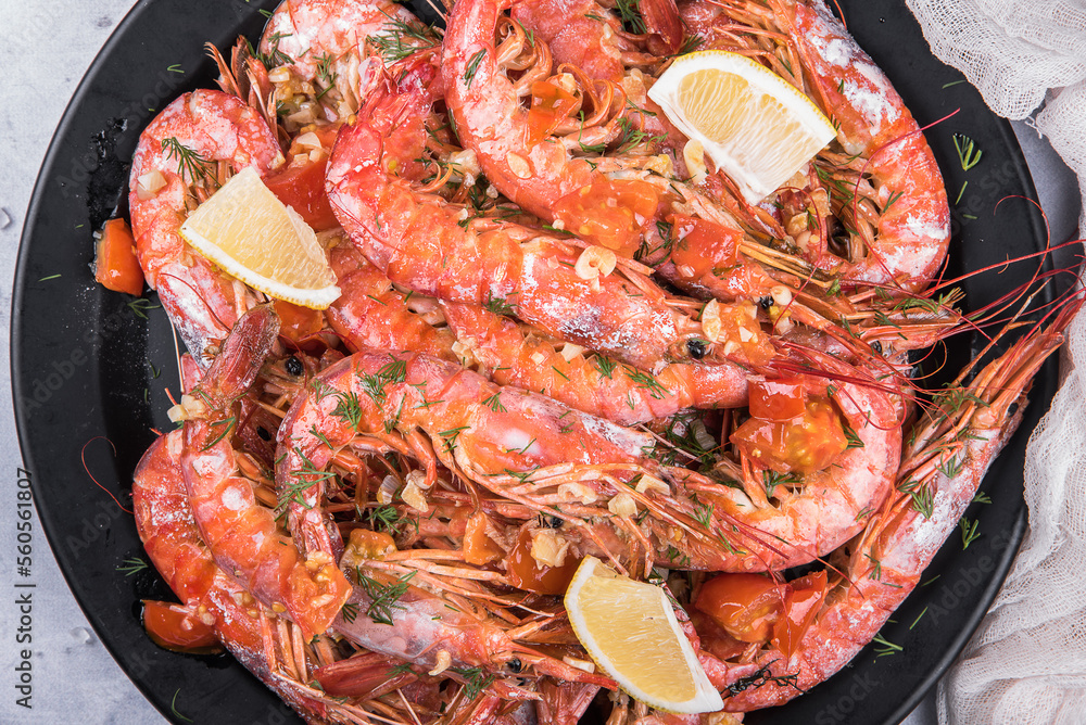 Baked langoustines in garlic sauce with cherry tomatoes on a black plate on a light marble background, top view. Baked langoustines in garlic sauce with cherry tomatoes on a black plate on a light marble background, top view.