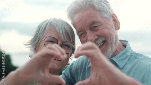 Close up portrait happy sincere middle aged elderly retired family couple making heart gesture with fingers, showing love or demonstrating sincere feelings together outdoors, looking at camera.